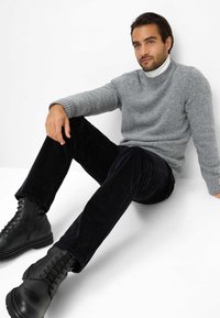 Grijze gebreide trui met ribbels aan de mouwen, zwarte corduroy broek en zwarte veterschoenen. Onderwerp zit tegen een witte achtergrond.