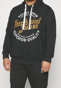 Svart sweatshirt med dragsko, med en stor grafik i orange och vit text. Ribbad mudd och nederkant, tillverkad av mjukt material.