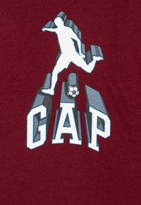 Vínová trička s bílou siluetou fotbalisty a slovy "GAP" ve stylizovaném modro-šedém písmu. Měkký bavlněný materiál.