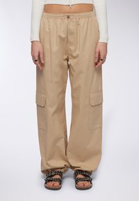Pantaloni cargo beige con una vestibilità ampia, elastico in vita, due grandi tasche laterali e una texture liscia. Abbinati a sandali neri con borchie.