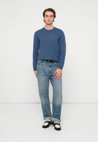 Niebieski sweter dziany, jasnoniebieskie dżinsy z podwiniętymi mankietami, czarny pasek i czarno-białe loafersy. Model stoi na tle białego tła.