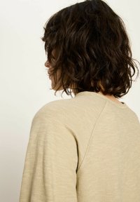 Frau mit schulterlangem, welligem, dunkelbraunem Haar, trägt ein beiges langärmliges Shirt, von der Seite und von hinten gegen einen schlichten Hintergrund betrachtet.