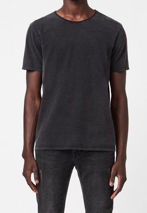 T-shirt noir à manches courtes, coupe ajustée, délavé, porté par une personne portant un jean slim délavé foncé, sur fond blanc.