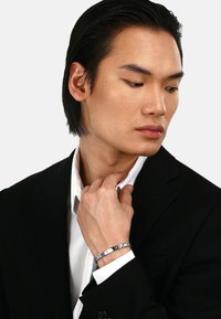 Silver bracelet med länkar med cirkulära och rektangulära accenter. Modell som bär en svart kostym och vit skjorta, som rör vid kragen med handen.