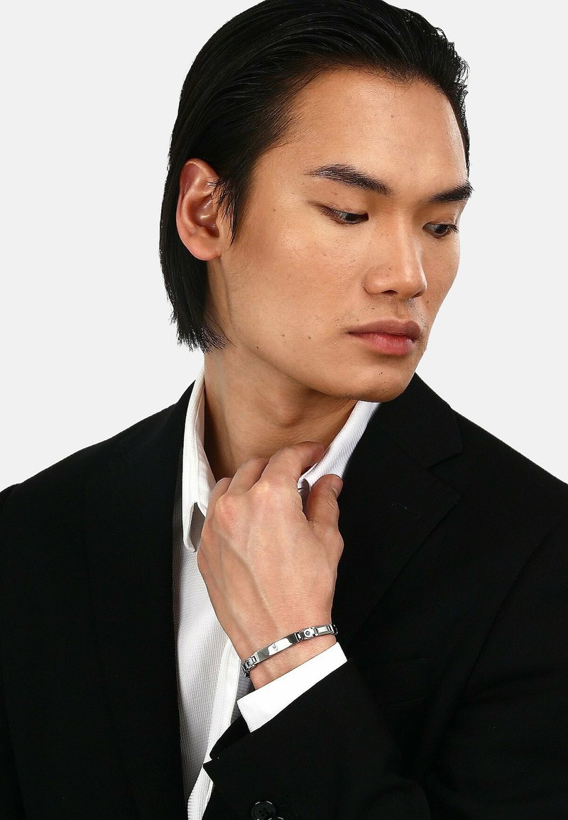 Silver bracelet med länkar med cirkulära och rektangulära accenter. Modell som bär en svart kostym och vit skjorta, som rör vid kragen med handen.