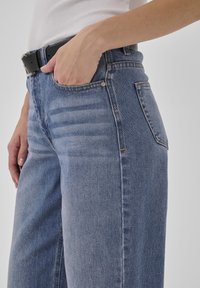 Oseba, ki nosi modre kavbojke iz jeansa s črnim pasom, roko v sprednjem žepu in vidno belo brezrokavnik z rebrasto teksturo.