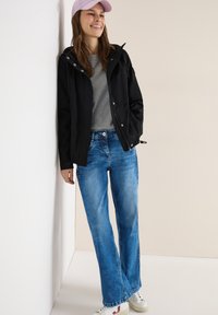 Femme portant une casquette rose, une veste à capuche noire, un t-shirt gris, un jean bleu et des baskets blanches, appuyée contre un mur blanc et souriante.