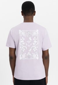 Lila katoenen t-shirt met een witte bloem- en vlinderafbeelding op de achterkant, omringd door een verfijnd randontwerp. Korte mouwen, ronde hals.