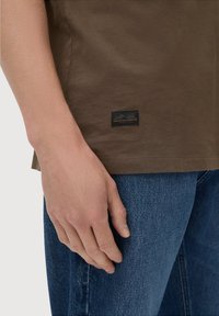Brun bomulds-t-shirt med rund halsudskæring, korte ærmer og et sort stofmærke ved kanten. Sat sammen med blå denim jeans.