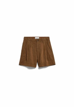 Vrouw staat, draagt een bruine mouwloze top en bijpassende korte broek met bruine en groene sandalen, tegen een effen witte achtergrond.