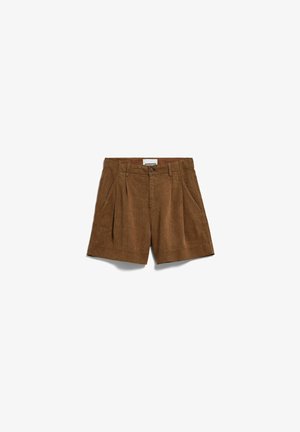 Vrouw staat, draagt een bruine mouwloze top en bijpassende korte broek met bruine en groene sandalen, tegen een effen witte achtergrond.