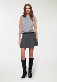 Maglione grigio senza maniche con motivo a trecce abbinato a una gonna a quadri nera e stivali alti in pelle nera, creando un look a strati.