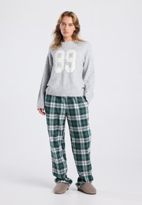 Jonge vrouw in een grijze trui met "89", groene geruite broek en beige slippers, staand tegen een effen lichte achtergrond.