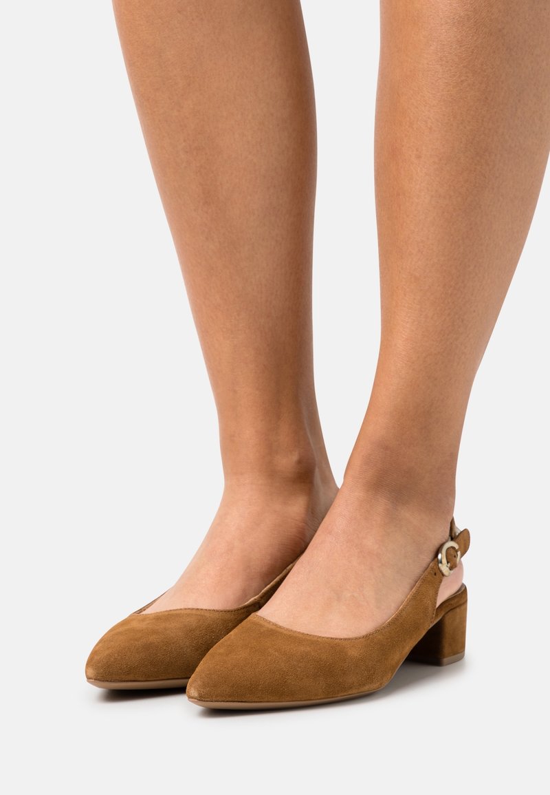 Jambes portant des chaussures slingback en daim marron avec des petits talons blocs et une petite boucle sur un fond clair.