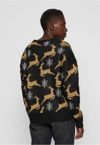 Pull noir en tricot avec motifs de rennes dorés et flocons de neige blancs. Coupe ample avec manches longues, poignets et ourlet côtelés.