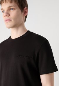 Juodas medvilnės marškinėliai su apvalia iškirpte ir išreikštu Calvin Klein logotipu ant krūtinės. Minkšta tekstūra, trumpomis rankovėmis ir klasikinio modelio.