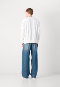 Sweatshirt bianco con maniche lunghe e polsini a coste, abbinato a jeans in denim blu a taglio largo con una texture sbiadita e leggeremente consumata.