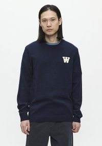 Pulover bleu marin cu guler rotund, manșete și tiv cu margini ondulate. Prezentă un logo texturat, de culoare crem, cu litera "W" pe pieptul stâng. Material moale.