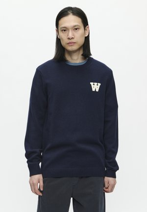 Navy blue sweater with a crew neck, ribbed cuffs, and hem. Features a textured, cream-colored "W" logo on the left chest. Soft material.