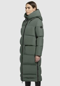 Giacca invernale lunga e imbottita di colore verde con colletto alto, chiusura a zip e tasche laterali; presenta pannelli trapuntati e un cordino sul cappuccio.