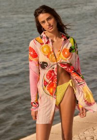 Multikleurige overhemd met fruitprints, met een lichtroze achtergrond en een open voorkant. Gecombineerd met gele gehaakte bikinislipjes.