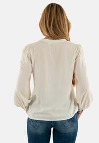 Blusa crema con mangas abullonadas, tejido texturizado y un sutil patrón de puntos. Acabado suave con un escote redondeado. Combinada con vaqueros azules.