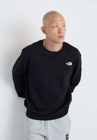 Svart genser med rund hals og subtil logo på brystet, kombinert med grå joggebukser. Glatt stoff og avslappet passform.