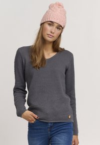 Grå teksturert genser med V-hals og lange ermer; brukt med blå jeans. Har en rund, trelogo detalj ved kanten.