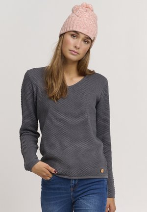 Grå tekstureret sweater med V-hals og lange ærmer; båret med blå jeans. Har en rund trælogo-detalje ved kanten.