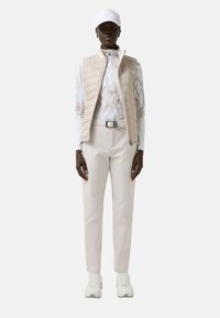 Béžová prošívaná vesta přes bílý top s dlouhým rukávem a vzorem, doplněná světlými kalhotami a bílými teniskami, k tomu bílá čepice.