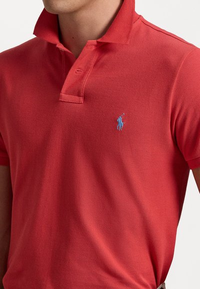 Punane polo särk, klassikalise kraega, lühikeste varrukatega ja õrna tekstuuriga kangast. Rinna peal asub sinine broderi logo.