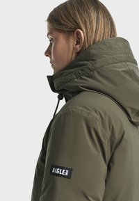 Olivovo zelená hybridná bunda s vysokým golierom, nastaviteľnou kapucňou a čiernym logom na rukáve, s prešívanou textúrou.