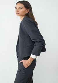 Donkergrijze cropped blazer met een enkele knoopsluiting, getailleerde pasvorm en opgerolde mouwen, gecombineerd met een wit shirt eronder.