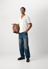 Lichte, gestructureerde crème blouse met golvenpatroon, blauwe jeans en zwarte schoenen. Houdt een bruin leren sporttas vast.