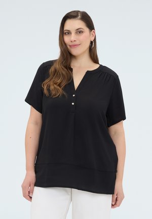 Frau trägt eine schwarze kurzärmelige Bluse mit Knöpfen und weiße Hose, steht vor einem schlichten hellen Hintergrund und lächelt leicht.