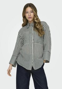 Camisa de botones a rayas en beige y verde oscuro. Cuenta con un collar, mangas largas y dos bolsillos en el pecho. Textura de tela suave y ligera.