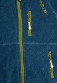 Giacca in pile blu con cerniere e cuciture verdi. Presenta due tasche con zip e una texture a coste. Logo "TROLLKIDS" ricamato in giallo.
