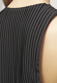 Gros plan sur l'épaule d'une personne portant un haut noir sans manches avec des plis verticaux espacés régulièrement.