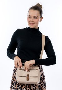 Frau trägt schwarzen Rollkragenpullover und Leopardenmuster-Rock, hält eine beigefarbene Umhängetasche mit braunen Schnallendetails und öffnet den Reißverschluss.