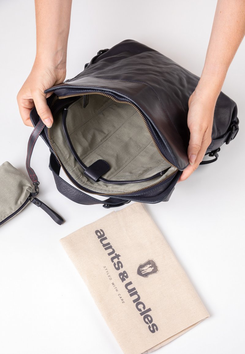 Zwarte leren rugzak met een ritssluiting. Het interieur heeft een gestructureerde beige voering en organisatiezakken. Inclusief een stoffen stofzak.