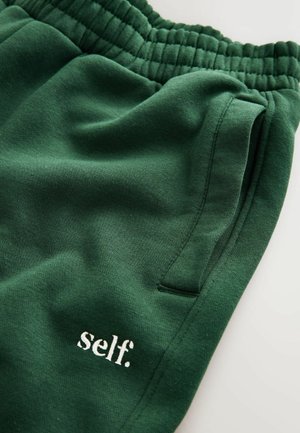 Groene joggingbroek van zachte stof, met een elastische tailleband, een zijzak en witte geborduurde tekst met de woorden "self."