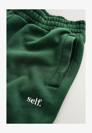 Green sweatpants made of soft fabric, featuring an elastic waistband, side pocket, and white embroidered text reading "self."