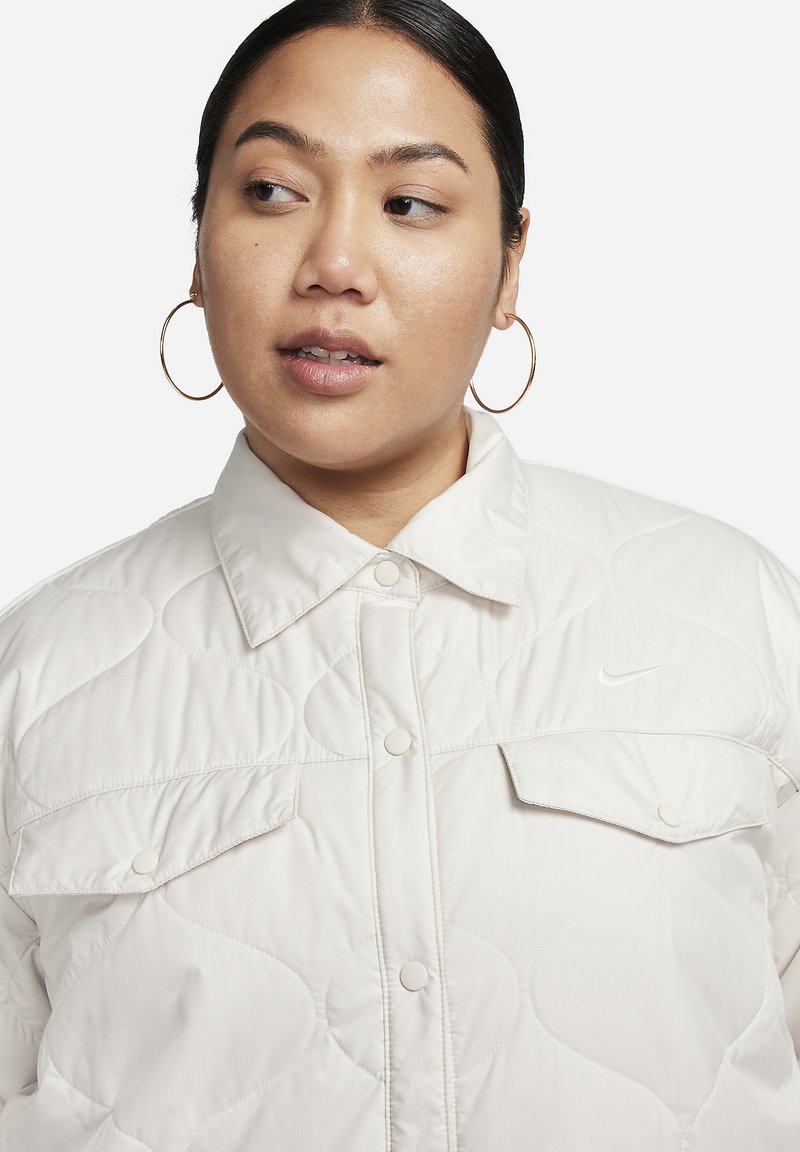 Veste matelassée crème avec des boutons-pression, deux poches poitrine et un logo Nike discret. Elle présente un col et une texture lisse et brillante.