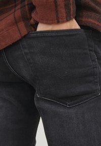 Pantalones vaqueros de mezclilla negra con un bolsillo trasero que presenta detalles de costura curva. La tela parece ligeramente texturizada, mostrando un diseño de corte clásico.