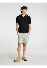 Črna polo majica s kratkimi rokavi in belimi detajli na ovratniku, v kombinaciji z zelenimi in belimi črtastimi hlačami, svetlimi sandali in rokami v žepih.