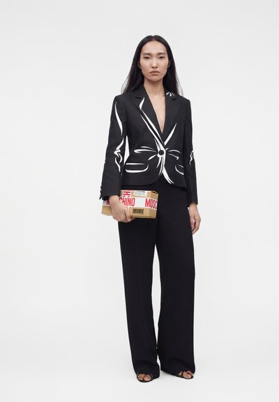 Femme en pantalon noir à jambes larges et blazer noir avec un imprimé abstrait blanc, tenant une petite boîte marron avec un ruban rouge Moschino.