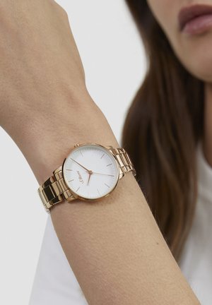 Montre ton or avec un cadran blanc rond, des index fins et un bracelet en métal maillé. Portée au poignet sur un fond neutre.