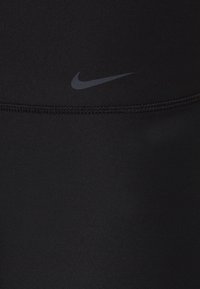 Musta urheiluhousut, jotka on valmistettu sileästä ja joustavasta kankaasta. Vyötärön lähellä on hillitty harmaa Nike-logo ja saumat on tikattu litteäksi.