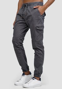 Graue Cargo-Hosen in lockerer Passform, mit Kordelzugbund, Seitentaschen und elastischen Bündchen, aus leichtem Baumwollstoff. Weiße Sneakers werden präsentiert.