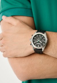 Chronografické hodinky s černým ciferníkem, stříbrným pouzdrem z nerezové oceli a černým gumovým řemínkem se zelenými akcenty a texturovanými detaily.